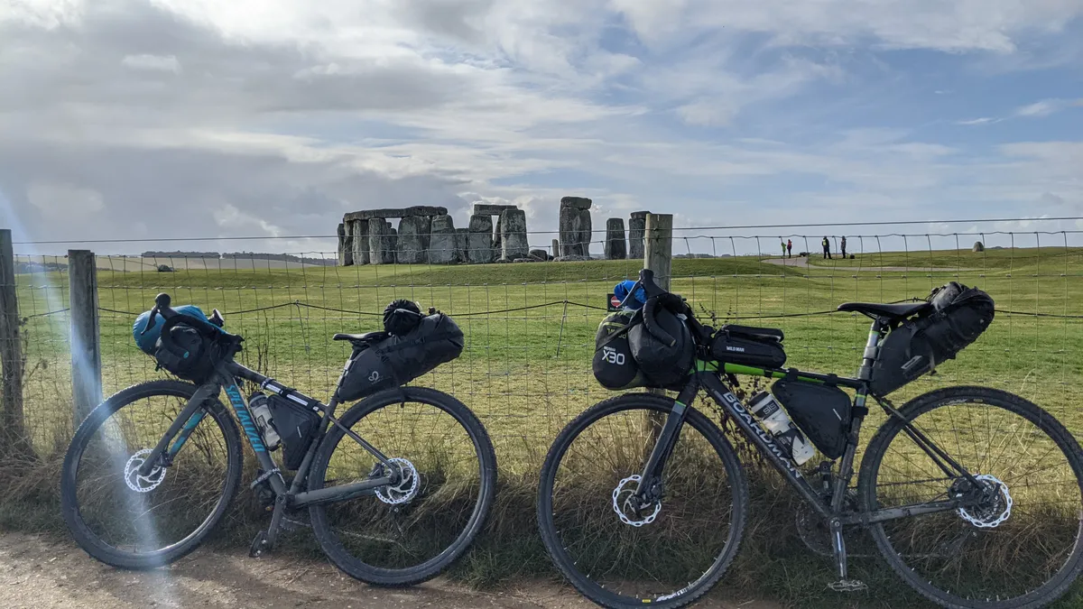 Two loaded bikepacking bikes leaning on a wire fence with Stonehenge behind them