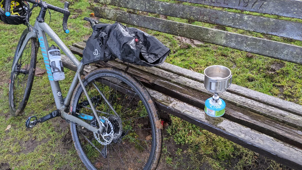 A muddy gravel bike next to a wet wooden bench with a tiny camping stove and cup brewing on it