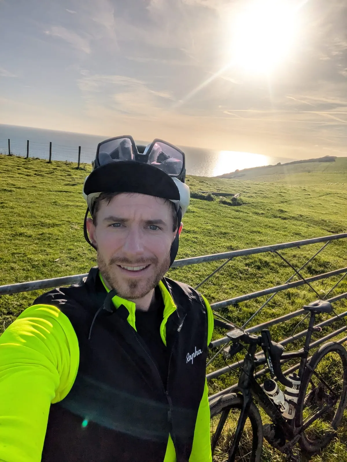 Selfie on a clifftop field above the sea with a muddy gravel bike leaning on a gate