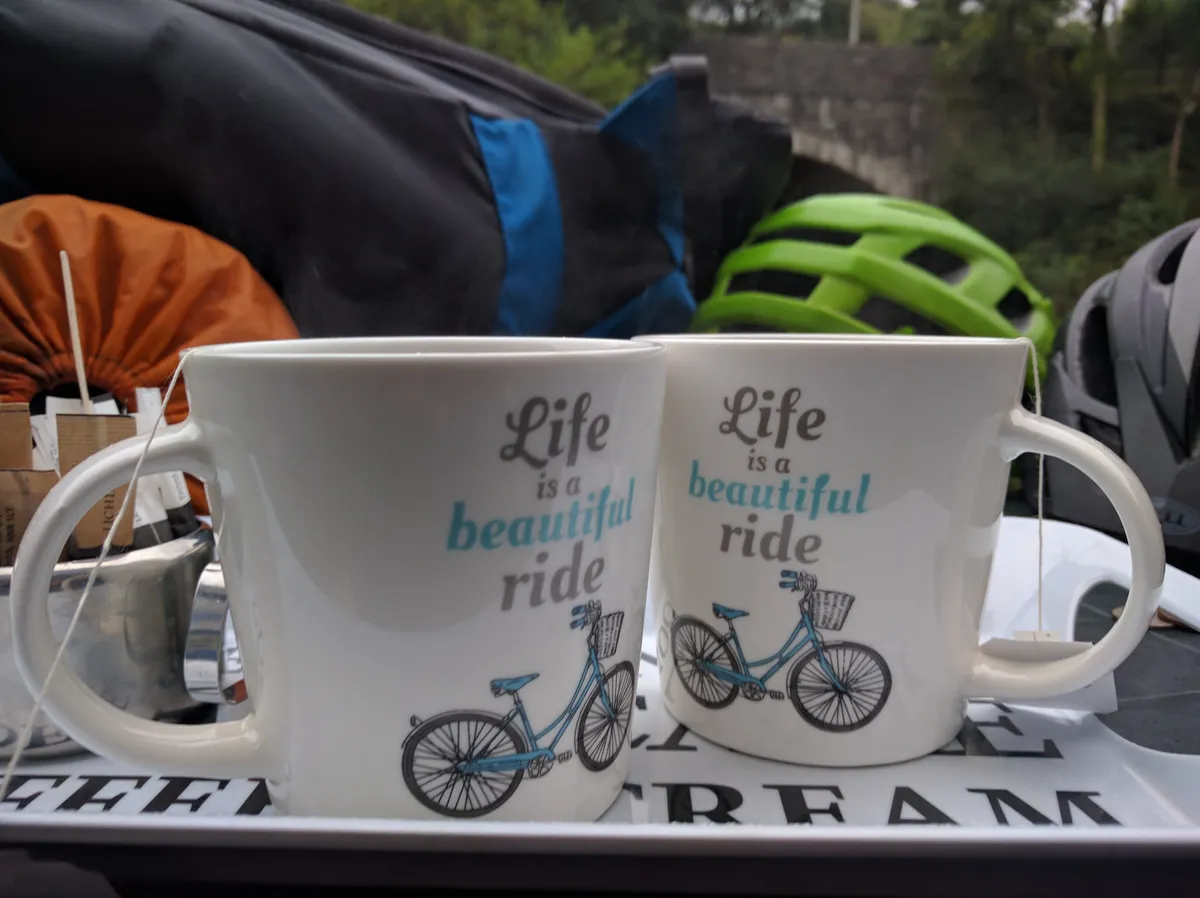 Two matching mugs reading 'Life is a beautiful ride' on a café tray, helmets and bags behind