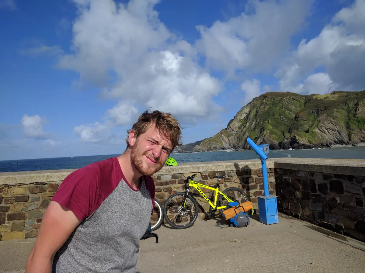 A younger brother squinting into the sun on a harbour wall, a loaded yellow hybrid bike and a green headland behind
