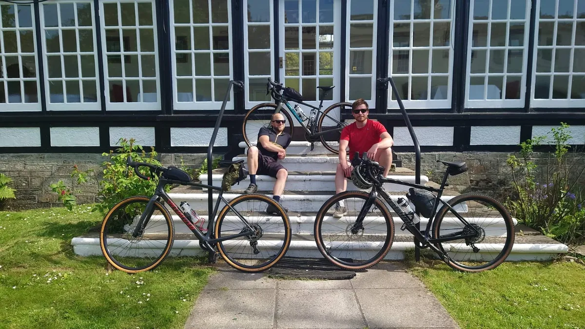 The brothers sitting on the steps of an old timber-framed building, gravel bikes propped on the lawn in front